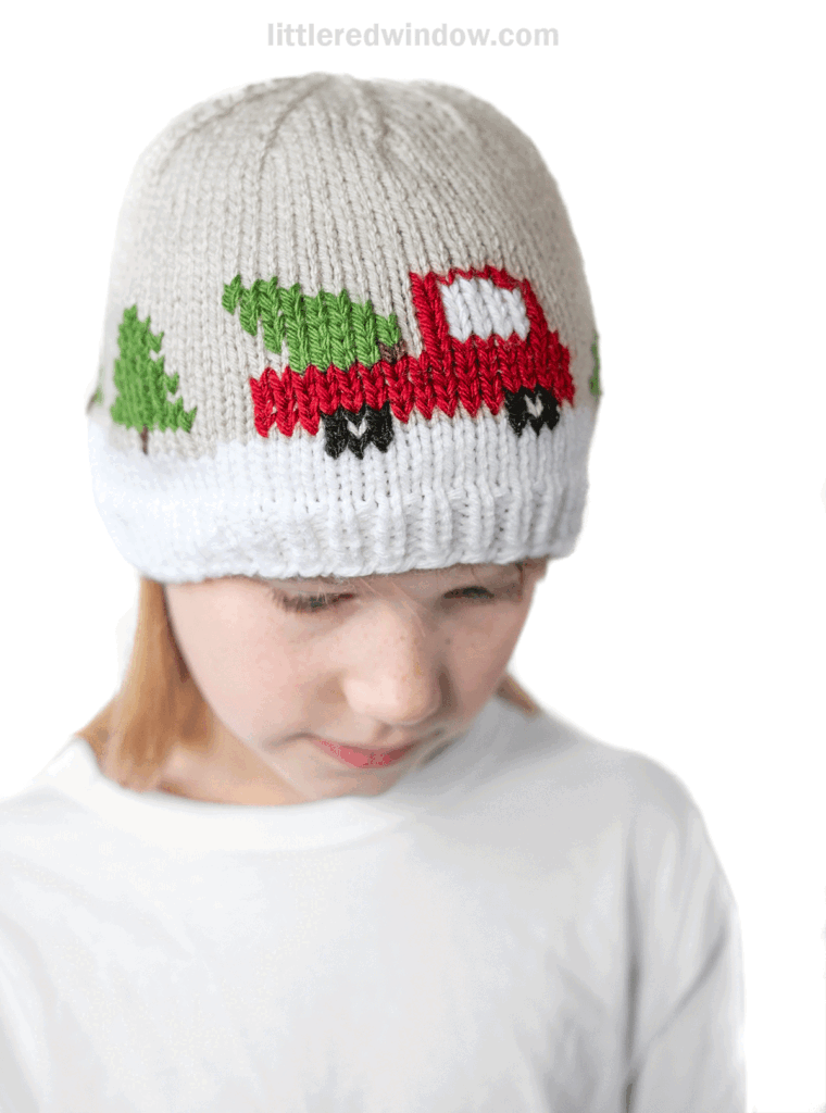 A child wearing a white and beige knit hat adorned with a christmas tree truck and festive green tree design, looks down while dressed in a plain white shirt.