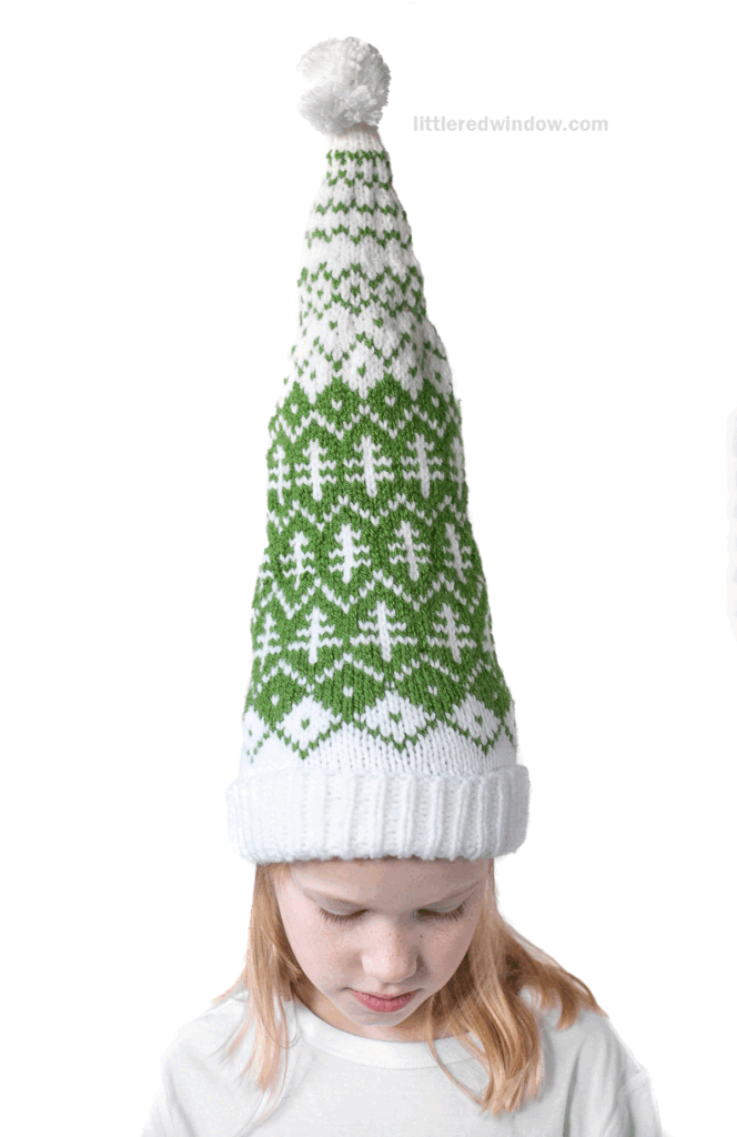 A young child wears an evergreen stocking cap with a white cuff, green and white geometric patterns, and a white pom-pom on top, looking down against a plain white background.