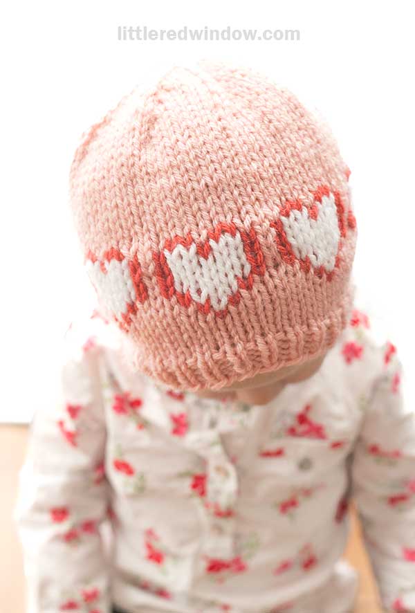A small child in a pink heart hat with white and red patterns sits wearing a matching floral top, set against a softly blurred background.