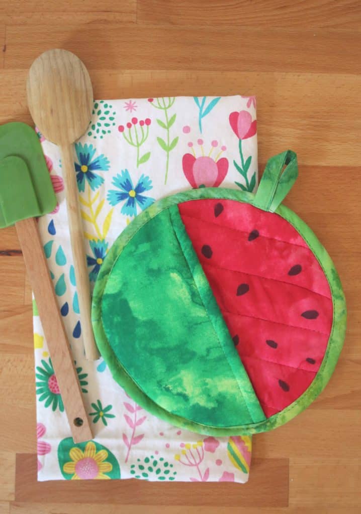 Round green potholder with one half painted red to look like watermelon on top of a tea towel and spatulas