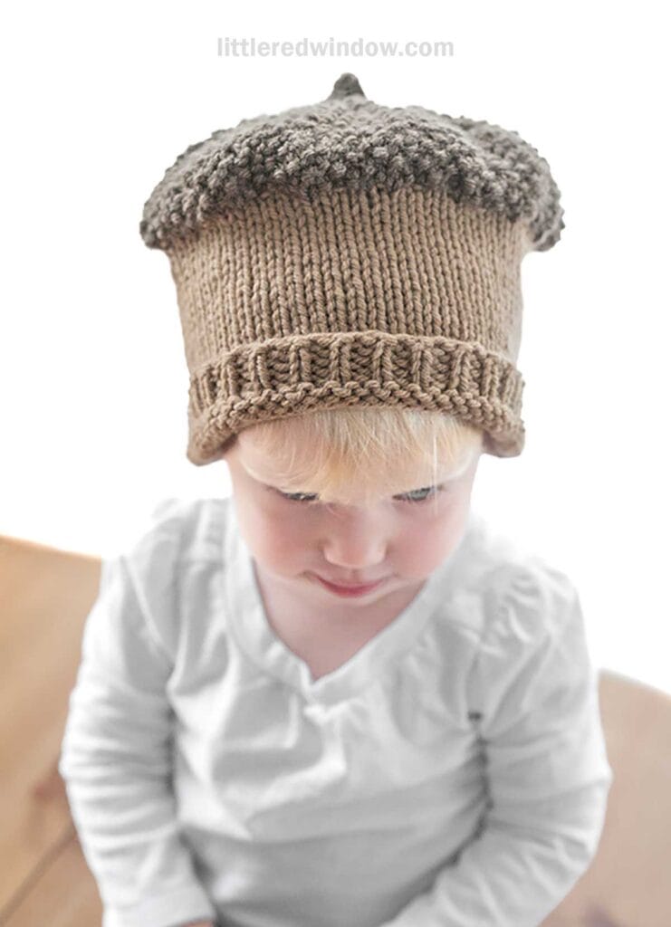 A young child with blonde hair gazes down, wearing an acorn hat that has a light brown body and a darker, textured top. Dressed in a white shirt, the child sits at a wooden surface.