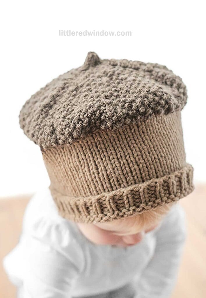 A child is wearing a light brown, knitted acorn hat with a textured top and ribbed brim, resembling an acorn cap. The hat has a small stem at the peak. The background is blurred, and the child is looking downwards.