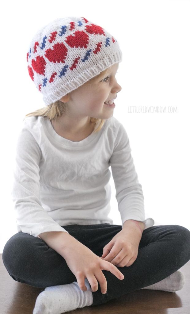 little girl sitting cross legged and wearing a white knit hat with red hearts and blue and red airmail stripes