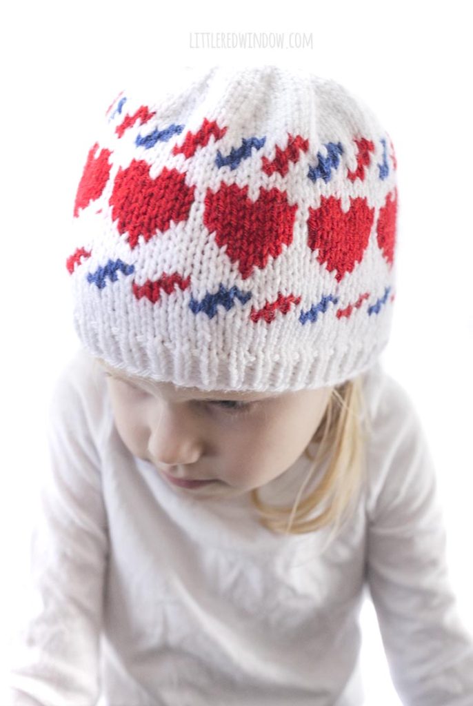 closeup of little girl and wearing a white knit hat with red hearts and blue and red airmail stripes