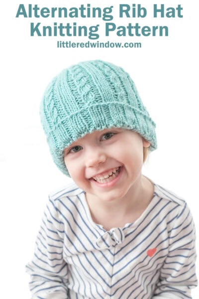 smiling girl in teal knit hat with alternating ribs of plain and cabled stitches and a folded up brim