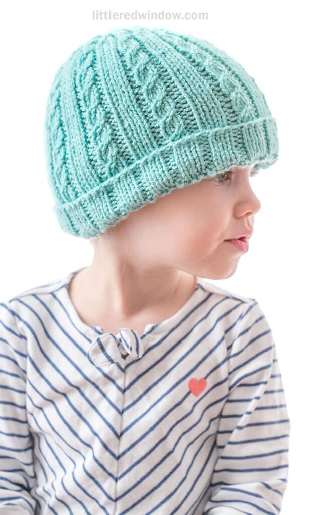 little girl in diagonal striped shirt wearing teal knit hat with alternating plain and cabled ribs looking off to the right