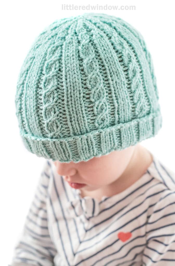 little girl in diagonal striped shirt wearing teal knit hat with alternating plain and cabled ribs looking down and to the left