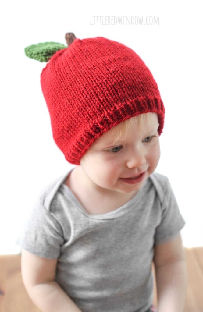 smiling baby in gray shirt wearing a red knit hat that looks like an apple with a brown stem and single green leaf on top sitting on a wood surface and looking slightly off to the right in front of a white background