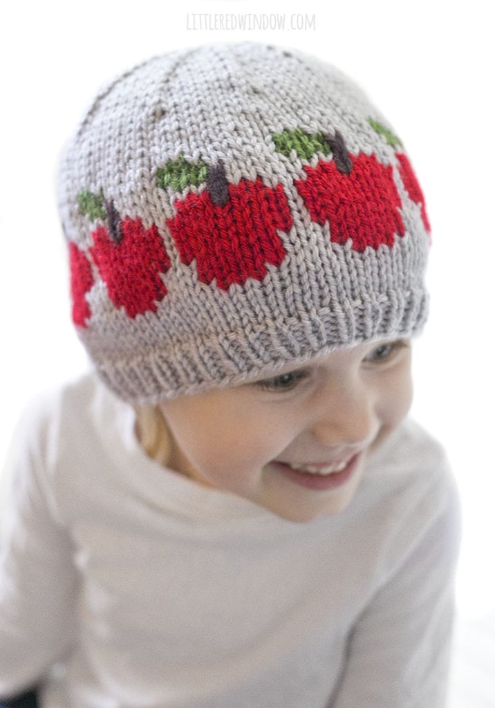 little girl smiling and wearing gray hat with red apples on it