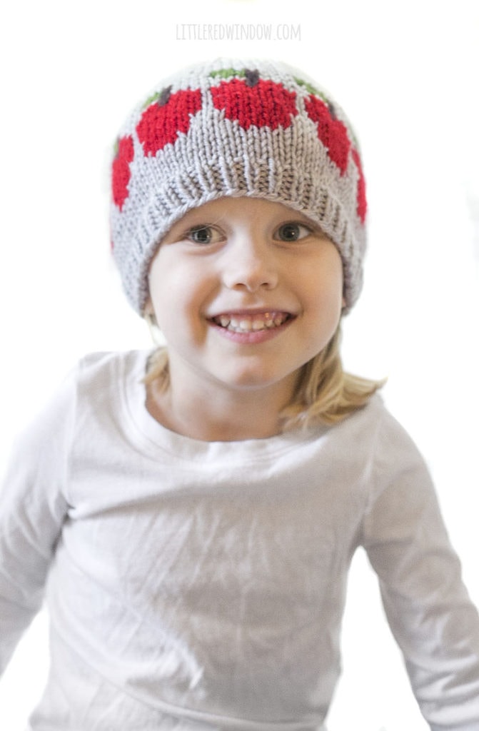 little girl in white shirt smiling and wearing gray knit hat with a row of red apples on it