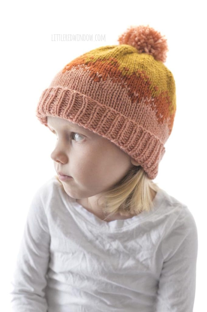 little blond girl in white shirt wearing pink rust and mustard colored autumn fade hat knitting pattern and looking off to the left
