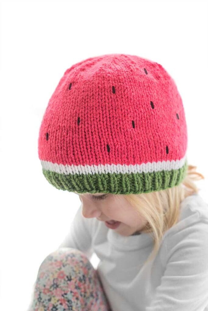 blonde child wearing a knit hat in green white and pink that looks like a watermelon slice looking down to the right