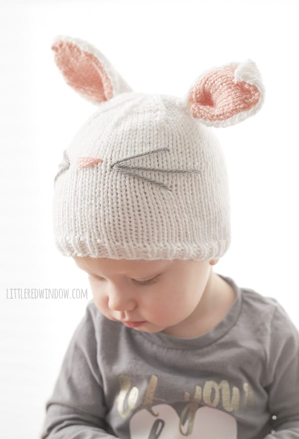 Chubby baby in gray shirt wearing a white knit hat that looks like a bunny and looking down to the left at their hands in front of a white background