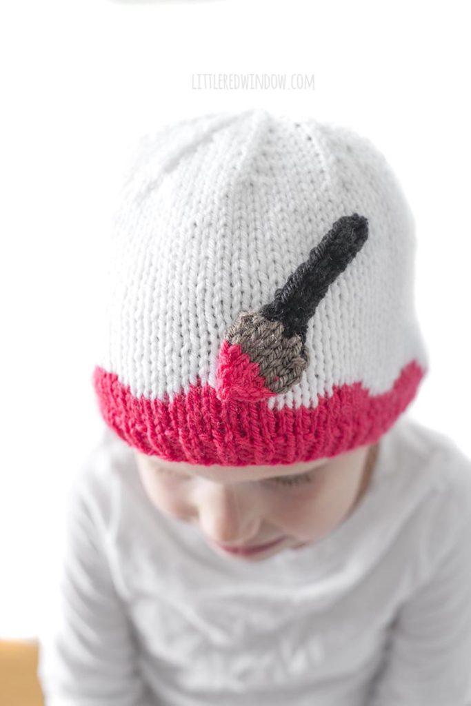 Little girl looking down and wearing a white hat with pink along the brim and an applique paintbrush painting on the pink