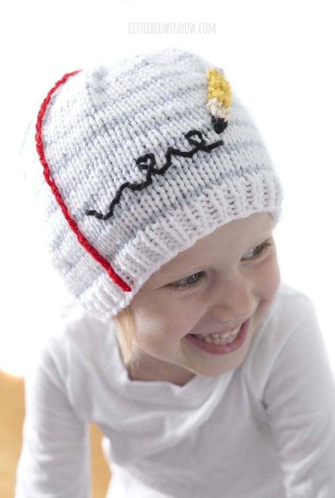 Little girl smiling and wearing a hat that looks like a piece of notebook paper with a pencil scribbling on it and looking to the right