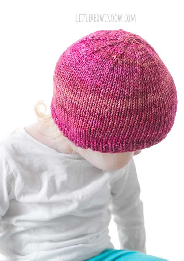 off center view of toddler looking down at their lap with one arm up in a white long sleeved tshirt in front of a white background wearing a basic knit hat with ribbed brim in ombre shades of raspberry and pink worsted weight yarn