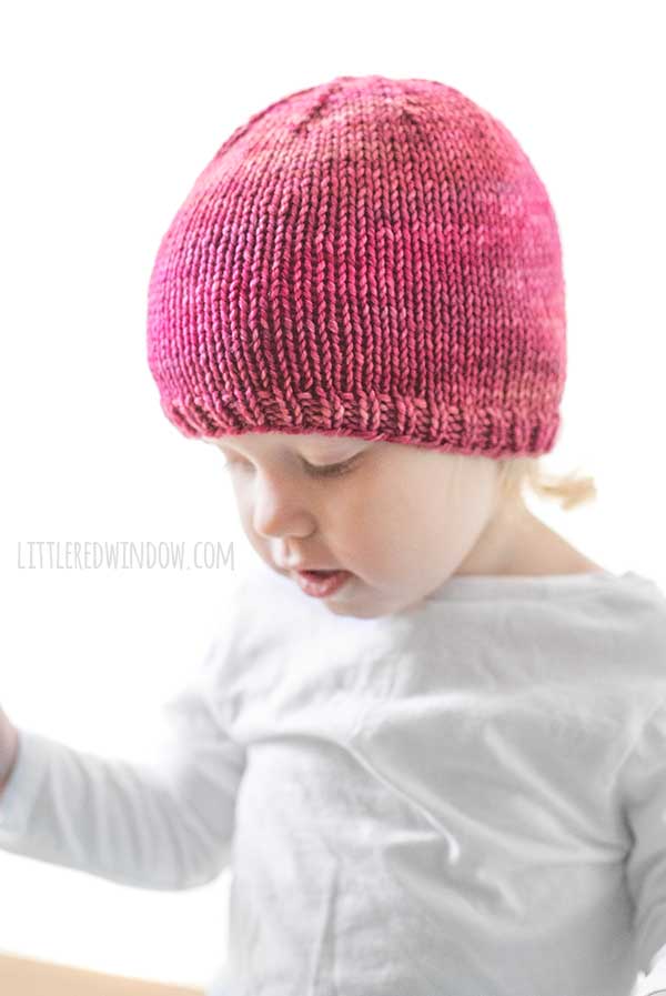 off center view of toddler looking down at their lap with one arm up in a white long sleeved tshirt in front of a white background wearing a basic knit hat with ribbed brim in ombre shades of raspberry and pink worsted weight yarn