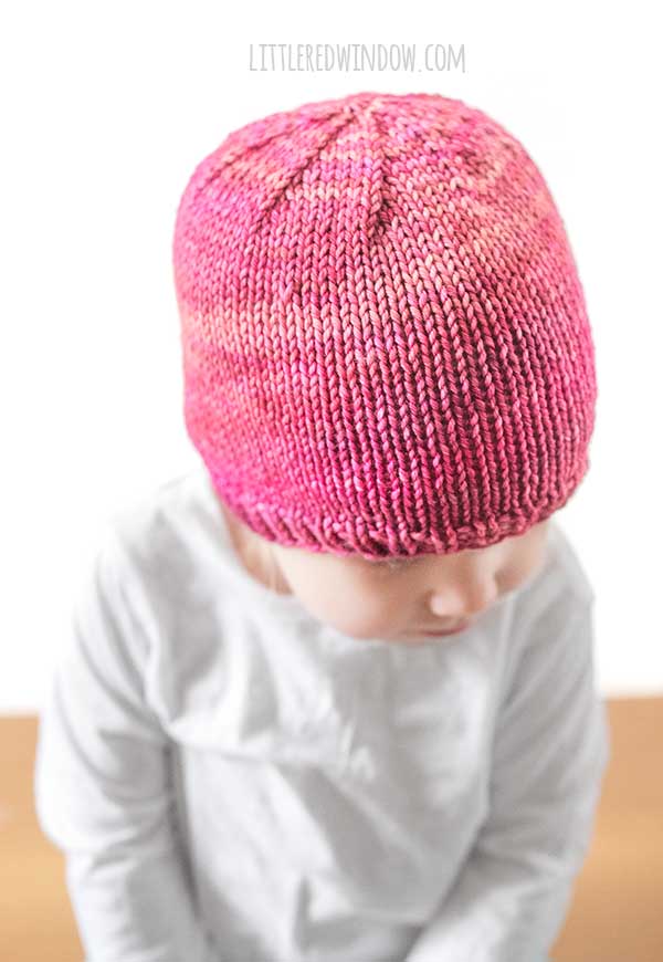 view of toddler looking down at their lap in a white long sleeved tshirt in front of a white background wearing a basic knit hat with ribbed brim in ombre shades of raspberry and pink worsted weight yarn