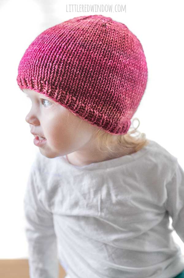 right side view of toddler in a white long sleeved tshirt in front of a white background wearing a basic knit hat with ribbed brim in ombre shades of raspberry and pink worsted weight yarn