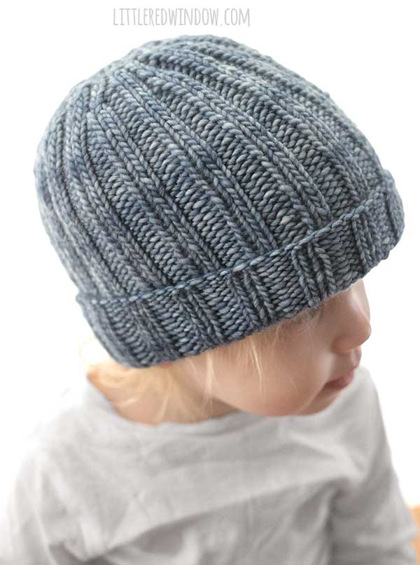 left side view of small child in gray shirt wearing a medium denim blue colored ribbed knit hat with folded up brim looking off into the distance in front of a white background