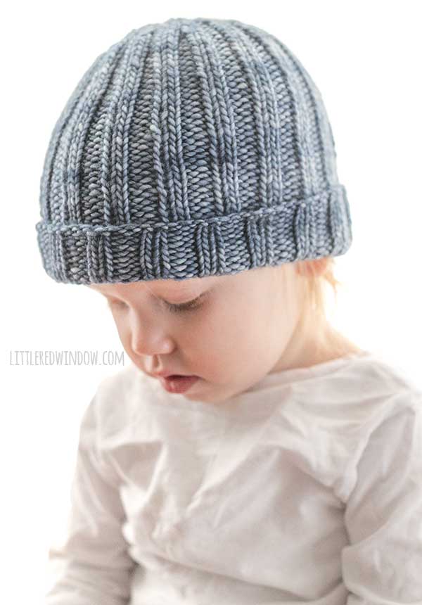 right side view of small child in gray shirt wearing a medium denim blue colored ribbed knit hat with folded up brim looking down at their lap in front of a white background