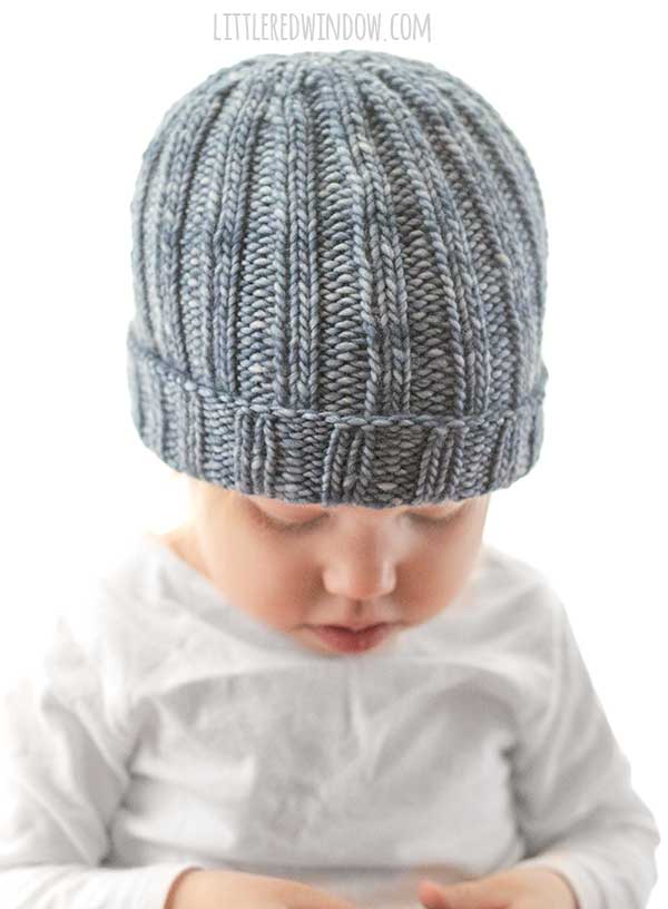 front side view of small child in gray shirt wearing a medium denim blue colored ribbed knit hat with folded up brim looking down at their hands in front of a white background