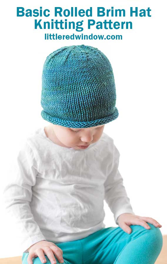 toddler in white shirt and blue pants wearing a knit hat with a rolled brim made of teal blue hand dyed yarn sitting on a wood table and looking down at their lap