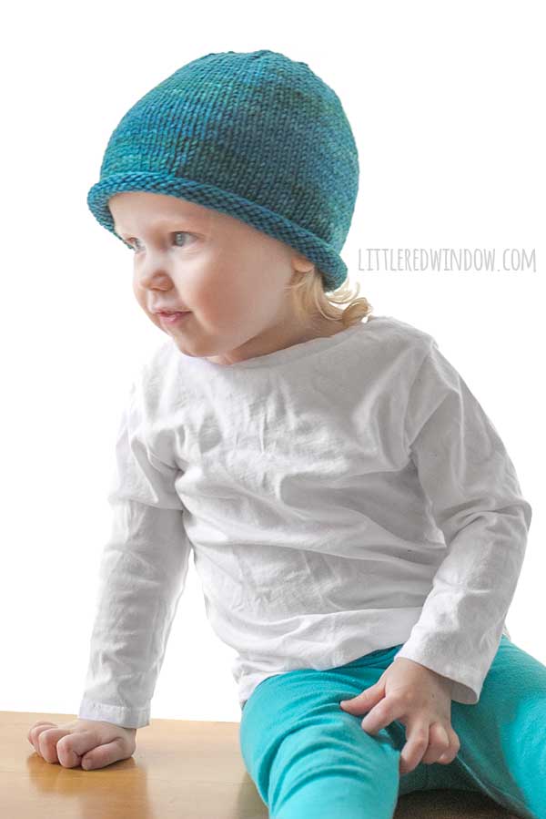 toddler in white shirt and blue pants wearing a knit hat with a rolled brim made of teal blue hand dyed yarn sitting on a wood table and leaning over to the left