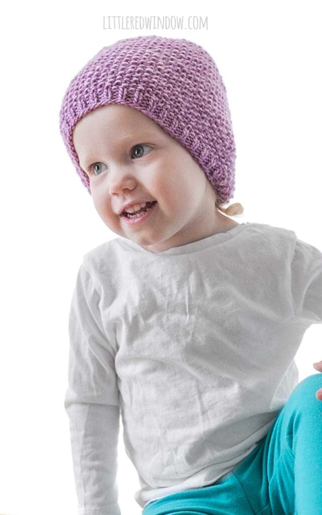 toddler in white shirt and teal pants leaning off to the left and wearing an orchid purple knit hat with small ribbed brim an the rest knit in seed stitch knitting pattern