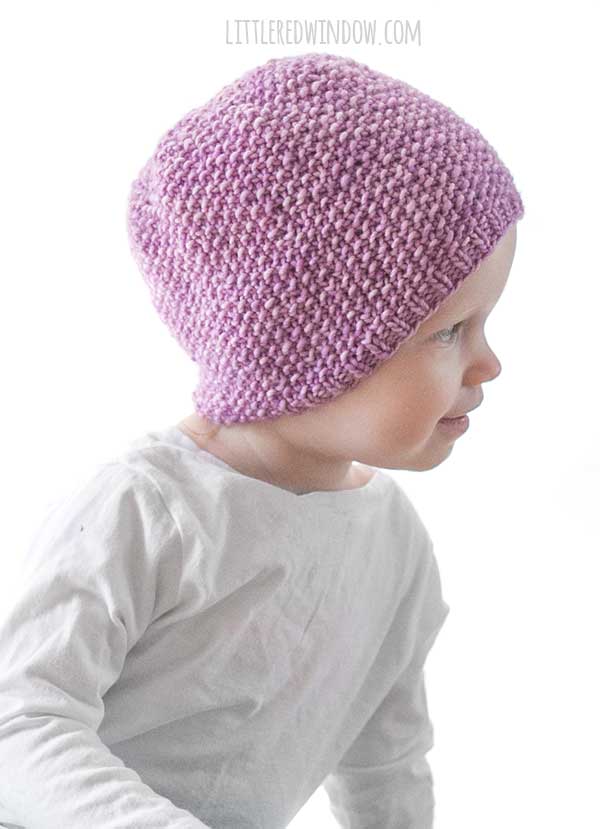 toddler in white shirt and teal pants leaning off to the left and wearing an orchid purple knit hat with small ribbed brim an the rest knit in seed stitch knitting pattern