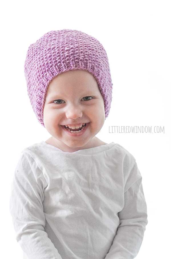 toddler in white shirt smiling at the camera wearing an orchid purple knit hat with small ribbed brim an the rest knit in seed stitch knitting pattern