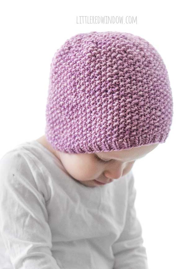 side view of toddler in white shirt looking down wearing an orchid purple knit hat with small ribbed brim an the rest knit in seed stitch knitting pattern