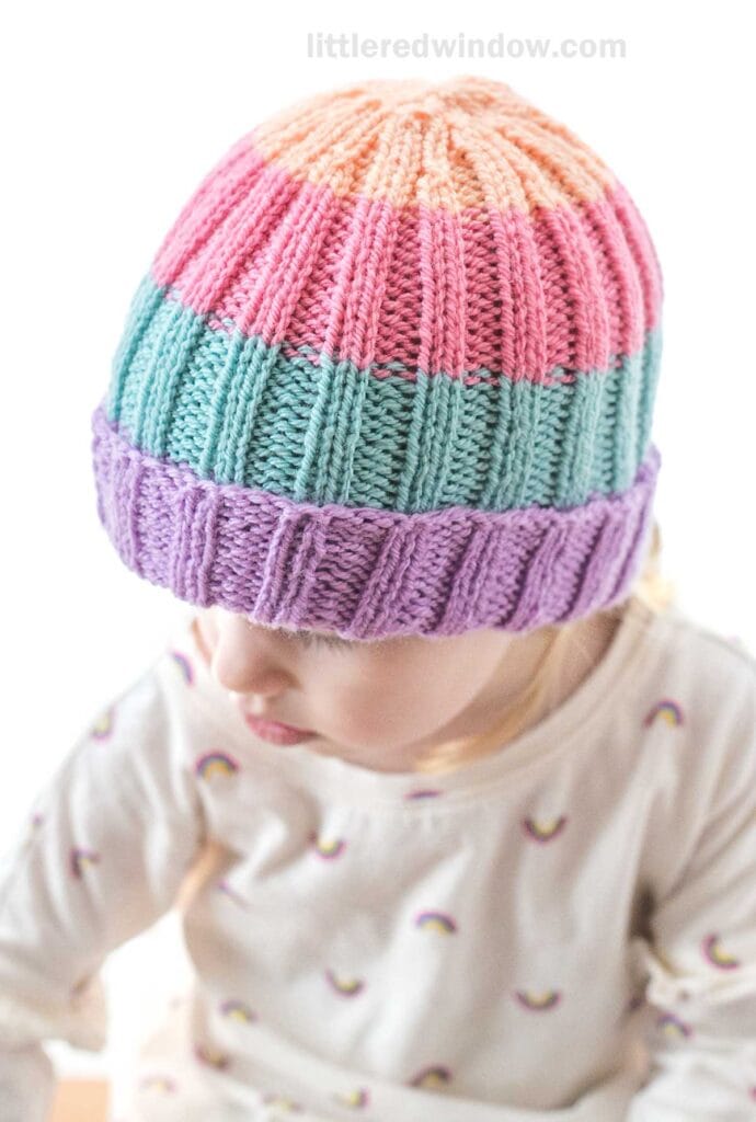 A child is wearing a knitted beanie with horizontal pastel-colored stripes in pink, peach, blue, and purple. The baby hat knitting pattern complements a light-colored top adorned with small rainbow patterns. The child is looking downward against a white background.