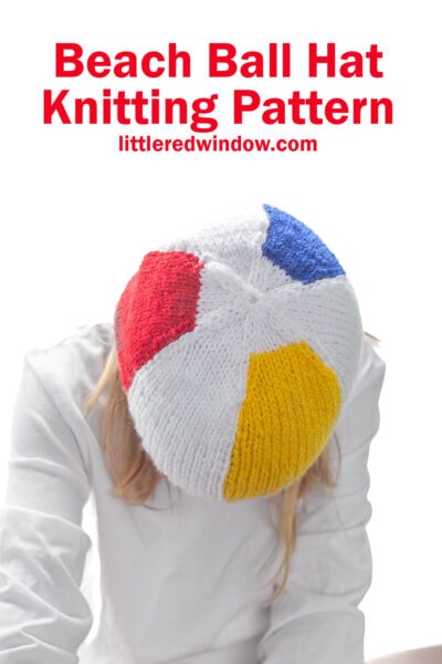child in white shirt leaning way forward to show the top of a knit beach ball hat