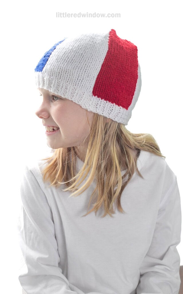 child in white shirt wearing a knit beach ball hat with white red and blue sections showing looking off to the left