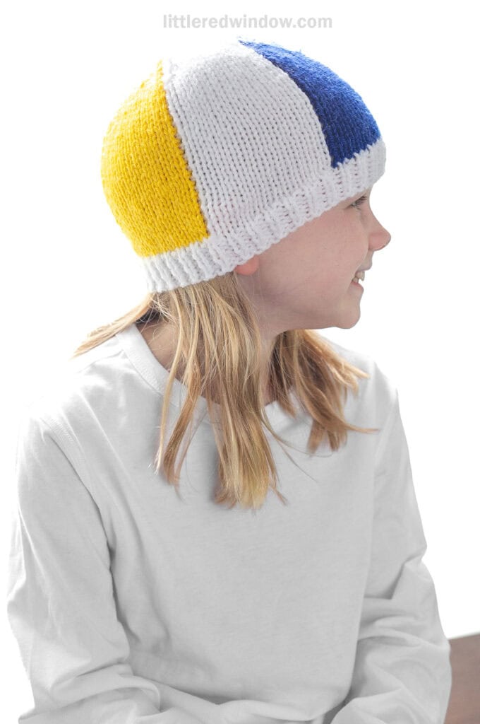 child in white shirt wearing a knit beach ball hat with white blue and yellow sections showing looking off to the right