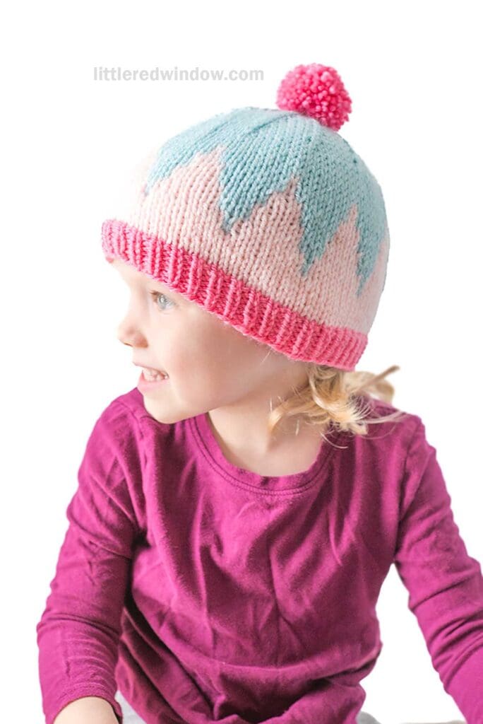 Young child in a bright pink shirt wearing a big zig hat—a knitted hat with a blue and pink zigzag pattern and a pink pom-pom—looking to the left against a white background.