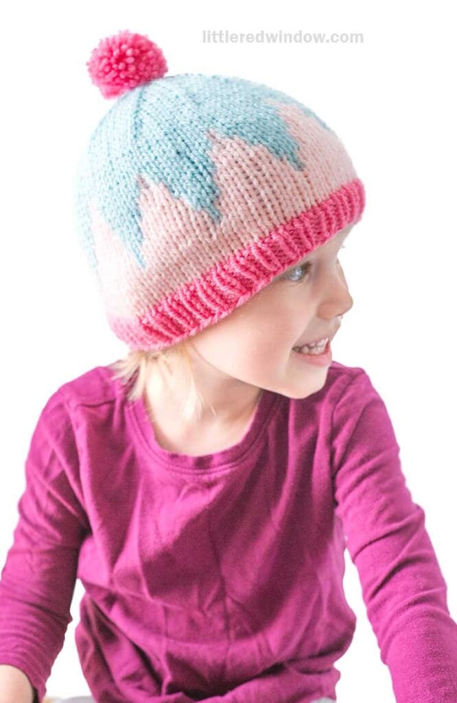 A young child wearing a pink long-sleeve shirt and a big zig hat with blue and pink zigzag patterns and a pink pom-pom, looking to the side and smiling against a white background.