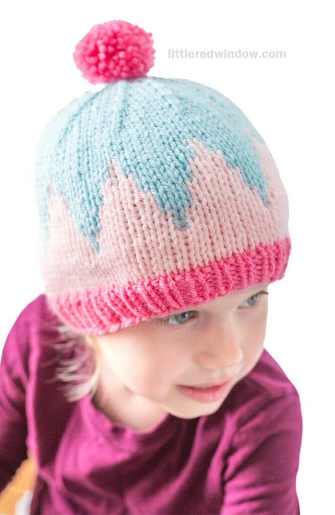 A young child wearing a big zig hat with a pink brim, light blue and white zigzag pattern, and a pink pom-pom on top. The child is dressed in a magenta shirt and looking to the side.
