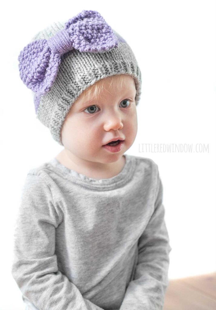 child wearing a gray shirt and gray knit hat with a purple band around the middle a large purple seed stitch bow on the front looking off in the distance to the near right