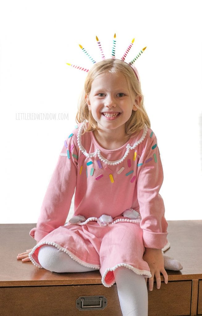 girl casually sitting on a table wearing a pink birthday cake costume with a candle headband