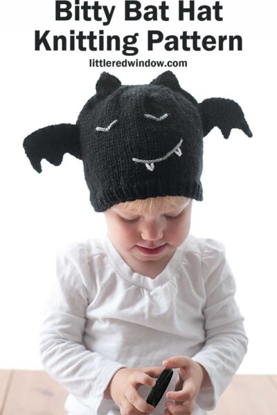 A young toddler wearing a charming black knit bat hat with wings, white eyes, and a smiling mouth with fangs holds an object while looking down. Text above reads "Bitty Bat Hat Knitting Pattern" and "littleredwindow.com".