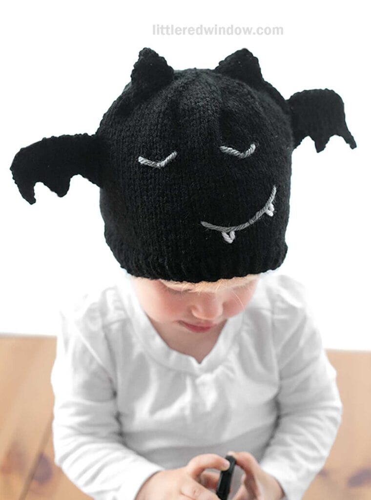 A young child is wearing a knitted black bat hat, complete with wings, eyes, and fangs. The child is dressed in a white long-sleeved shirt and is looking down while holding an object. The background is neutral.
