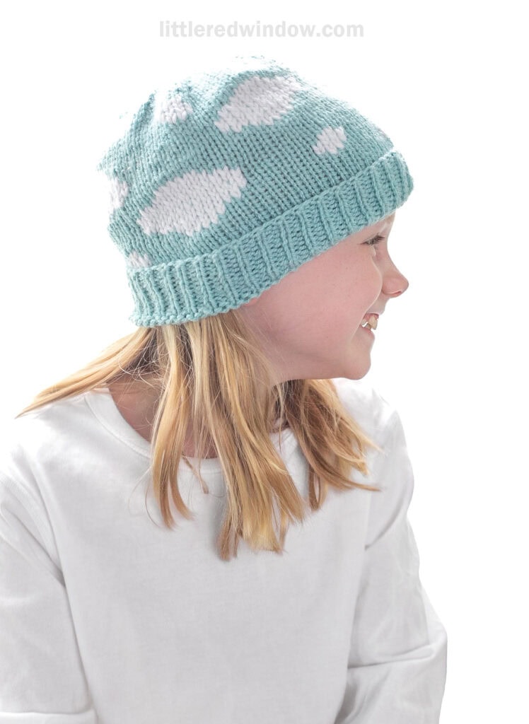 girl in white shirt wearing a light blue knit hat with ribbed brim covered in a white cloud pattern looking off to the right and smiling in front of a white background