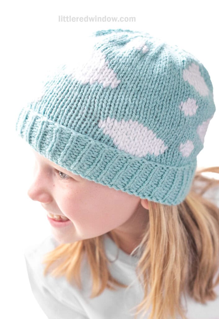 view from slightly above of child wearing blue sky knit hat with white clouds all over it looking off to the left and smiling