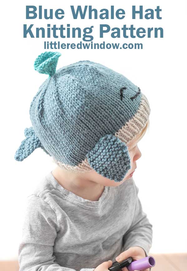 Toddler in a gray shirt wearing a blue knit whale hat in front of a white background looking off to the right