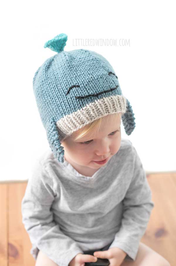 Toddler in a gray shirt wearing a blue knit whale hat in front of a white background looking down and to the right