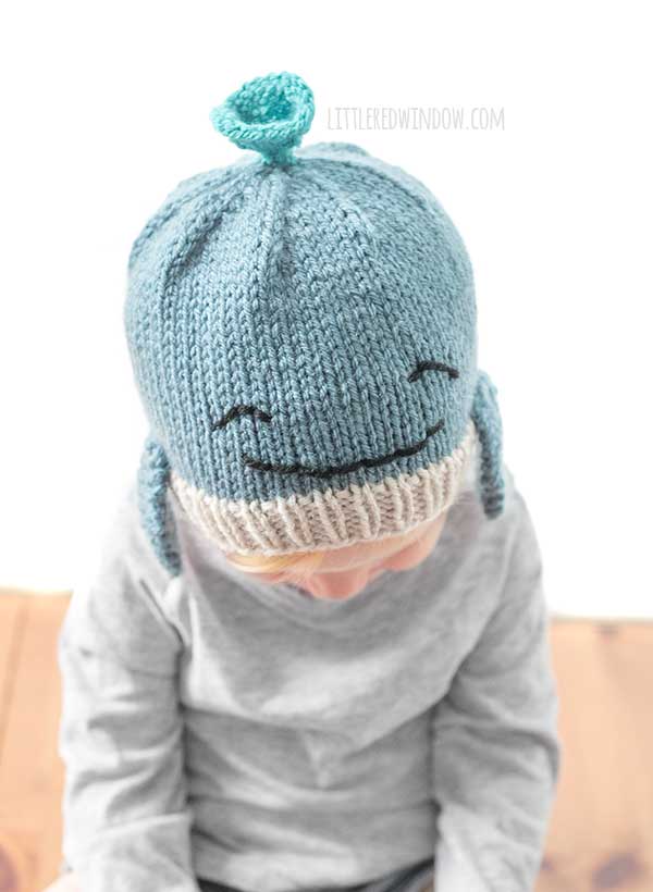 Toddler in a gray shirt wearing a blue knit whale hat in front of a white background looking down at their lap
