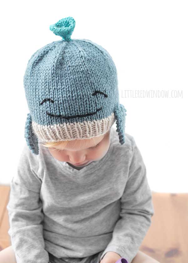 Toddler in gray shirt wearing a blue knit whale hat with smiling face on it looking down at something in their hands
