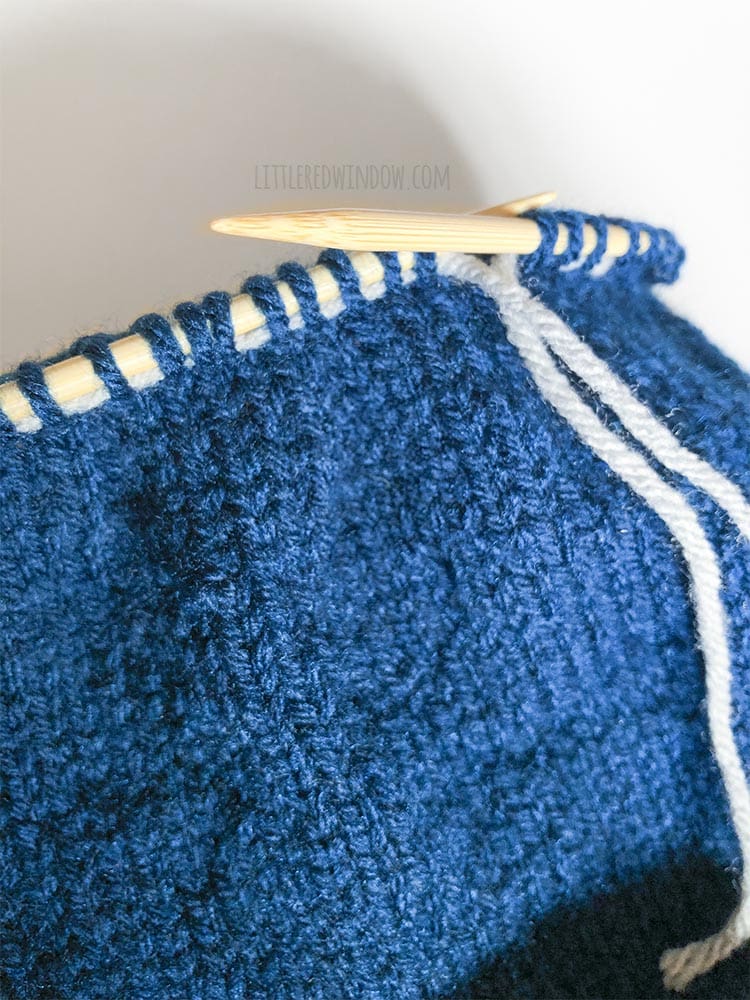 closeup of a blue knit hat being knit with a lifeline inserted through the live knitting stitches